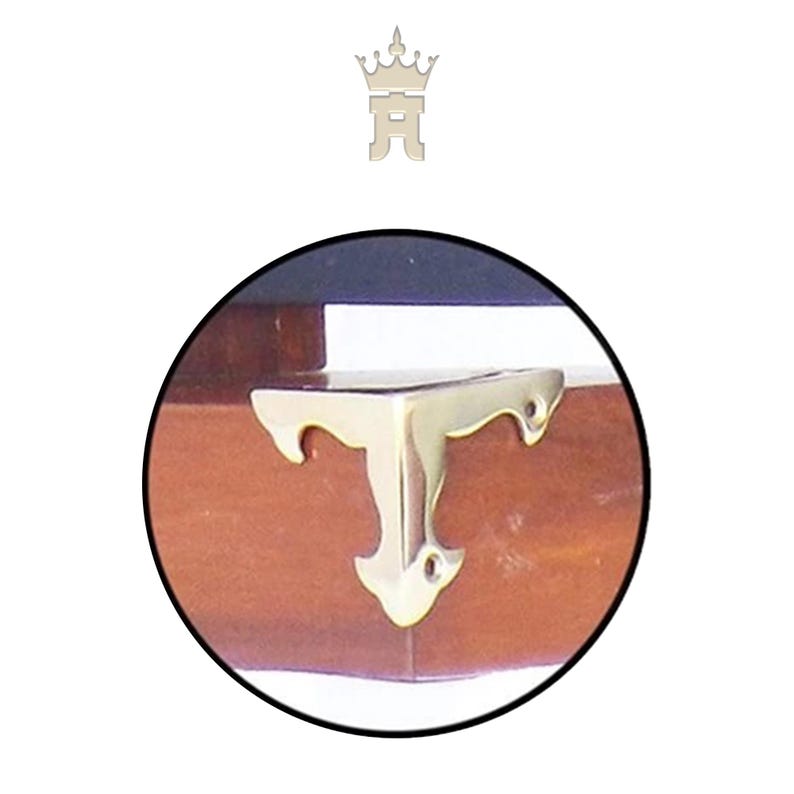 Puede incluir: Primer plano de un soporte de esquina de lat&oacute;n fijado a una superficie de madera. El soporte tiene forma de T con curvas decorativas y peque&ntilde;os agujeros para tornillos. La madera es de color marr&oacute;n c&aacute;lido.