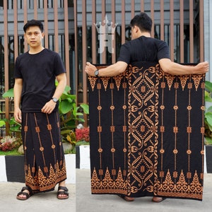 Puede incluir: Un sarong negro con un diseño estampado en marrón y dorado. El sarong lo lleva puesto una persona de pie delante de una valla de madera.