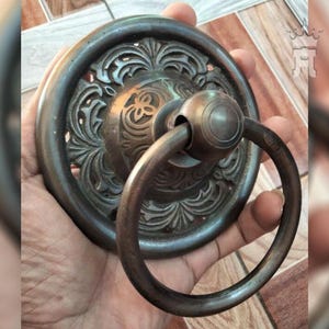 Könnte beinhalten: Ein großer, verzierter, antiker Türklopfer aus Messing mit einem Ringgriff. Der Türklopfer hat ein kreisförmiges Design mit komplizierten floralen Mustern.