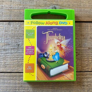 Puede incluir: Una caja de DVD verde con el título "Thumbelina". La caja tiene un asa y texto que dice "Follow Along DVD", "Full Length Movie" y "Kids Captioning In English And Spanish". La portada presenta una escena de cuento de hadas.