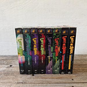 May include: A collection of eight VHS tapes of the television show "Lost in Space." The tapes have colorful covers with the show's logo and episode titles. The tapes are arranged vertically on a wooden surface.