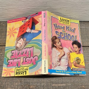 Puede incluir: Dos libros de bolsillo con cubiertas rosa y amarilla que muestran a una joven en la portada. El libro de la izquierda se titula "Just Like Lizzie" y el libro de la derecha se titula "New Kid in School" con el texto "Lizzie McGuire" sobre el título. Ambos libros son de la exitosa serie de televisión de Disney Channel.