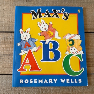 May include: A children's book titled "Max's ABC" by Rosemary Wells. The cover features a blue border with a yellow center. The title is in large, bold letters, with illustrations of rabbits interacting with the letters A, B, and C.