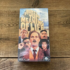 May include: A VHS tape case for the movie Monty Python's The Meaning of Life. The case features a collage of the main actors from the film, including John Cleese, Michael Palin, and Terry Gilliam. The text "Monty Python's The Meaning of Life" is printed in large, bold letters on the front of the case.