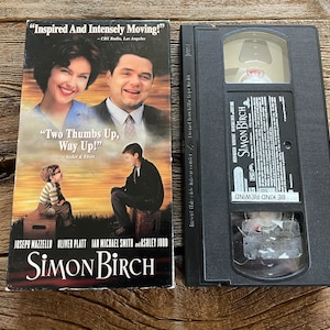 May include: VHS video cassette tape of the movie "Simon Birch" featuring a photo of the actors on the front cover. The cover text reads "Inspired And Intensely Moving!" and "Two Thumbs Up, Way Up!"