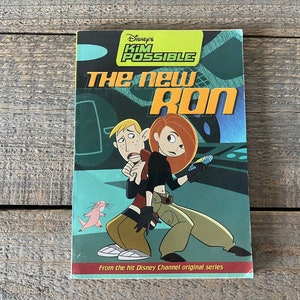 以下が含まれることがあります： ディズニーの「キム・ポッシブル」のDVDケースで、「The New Ron」というタイトルです。カバーアートは、キム・ポッシブルとロン・ストッパブルがアクションポーズをとっています。背景はティール、グリーン、ブルーのミックスです。「From the hit Disney Channel original series」の文字が下部にあります。