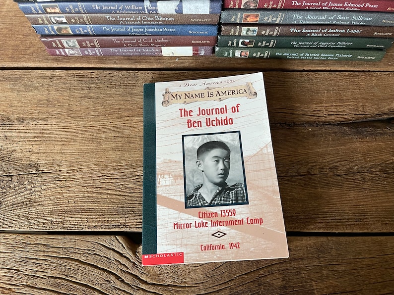 Puede incluir: Un libro de bolsillo titulado "My Name Is America: The Journal of Ben Uchida" con una foto de un ni&ntilde;o peque&ntilde;o en la portada. El libro forma parte de la serie "Dear America" y cuenta la historia de un ni&ntilde;o japon&eacute;s-estadounidense que fue internado durante la Segunda Guerra Mundial. La portada del libro tambi&eacute;n incluye el texto "Citizen 13559, Mirror Lake Internment Camp, California, 1942."