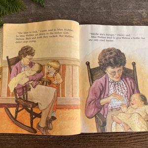 Pu&ograve; includere: Un libro aperto a due pagine con illustrazioni di persone che cullano un bambino e che lo nutrono con un biberon. Il testo sulla pagina dice "Le piace dondolarsi", disse Carrie a Miss Webber. Cos&igrave; Miss Webber si sedette nella sedia a dondolo con Melissa. Si dondolavano avanti e indietro. Ma Melissa non smise di piangere. "Forse ha fame", disse Davey. Miss Webber cerc&ograve; di dare un biberon a Melissa, ma lei piangeva solo pi&ugrave; forte."