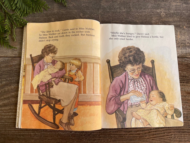 Pu&ograve; includere: Un libro aperto a due pagine con illustrazioni di persone che cullano un bambino e che lo nutrono con un biberon. Il testo sulla pagina dice "Le piace dondolarsi", disse Carrie a Miss Webber. Cos&igrave; Miss Webber si sedette nella sedia a dondolo con Melissa. Si dondolavano avanti e indietro. Ma Melissa non smise di piangere. "Forse ha fame", disse Davey. Miss Webber cerc&ograve; di dare un biberon a Melissa, ma lei piangeva solo pi&ugrave; forte."