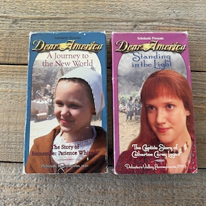 Puede incluir: Dos cintas VHS de la serie Dear America. La cinta de la izquierda se titula "A Journey to the New World" y presenta a una joven con un gorro blanco. La cinta de la derecha se titula "Standing in the Light" y presenta a una joven con el pelo largo y rojo.