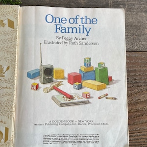Pu&ograve; includere: Un libro vintage Golden Book intitolato "One of the Family" di Peggy Archer, illustrato da Ruth Sanderson. La copertina del libro presenta un'illustrazione colorata di un bambino che gioca con blocchi di legno e una radio giocattolo.