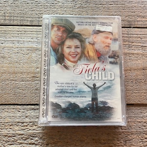 Puede incluir: Caja de DVD con una imagen de tres personas en la parte delantera. El título de la película es "Fiela's Child". El texto en la caja dice "La épica historia del amor de una madre por su hijo adoptivo y el hombre en que se convirtió. Un drama humano cargado de emociones."