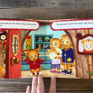 Puede incluir: Una p&aacute;gina doble de un libro infantil con personajes de Daniel el Tigre. Una p&aacute;gina muestra a Daniel con una sudadera roja, el texto dice: "&iexcl;Daniel tiene noticias geniales que compartir. &iexcl;Es un hermano mayor!" La otra p&aacute;gina muestra a la familia de Daniel con un beb&eacute;.