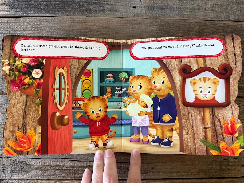 Puede incluir: Una p&aacute;gina doble de un libro infantil con personajes de Daniel el Tigre. Una p&aacute;gina muestra a Daniel con una sudadera roja, el texto dice: "&iexcl;Daniel tiene noticias geniales que compartir. &iexcl;Es un hermano mayor!" La otra p&aacute;gina muestra a la familia de Daniel con un beb&eacute;.