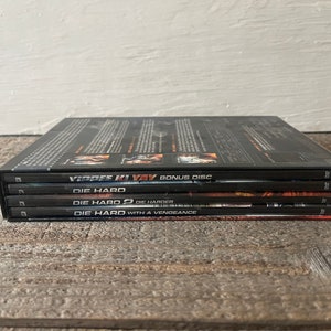 K&ouml;nnte beinhalten: Ein Stapel von vier DVD-H&uuml;llen f&uuml;r die Die Hard-Filmreihe. Die oberste H&uuml;lle ist eine Bonus-Disc f&uuml;r den Film "Stirb Langsam 4.0". Die anderen H&uuml;llen sind f&uuml;r "Stirb Langsam", "Stirb Langsam 2: Stirb Langsamer", und "Stirb Langsam: Jetzt erst recht".