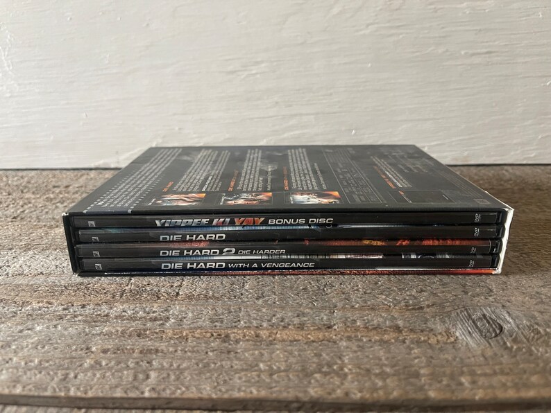 K&ouml;nnte beinhalten: Ein Stapel von vier DVD-H&uuml;llen f&uuml;r die Die Hard-Filmreihe. Die oberste H&uuml;lle ist eine Bonus-Disc f&uuml;r den Film "Stirb Langsam 4.0". Die anderen H&uuml;llen sind f&uuml;r "Stirb Langsam", "Stirb Langsam 2: Stirb Langsamer", und "Stirb Langsam: Jetzt erst recht".