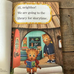 May include: A children's book page with the text "Hi, neighbor! We are going to the library for storytime." The illustration features a tiger character holding a book and another tiger character standing in a doorway.