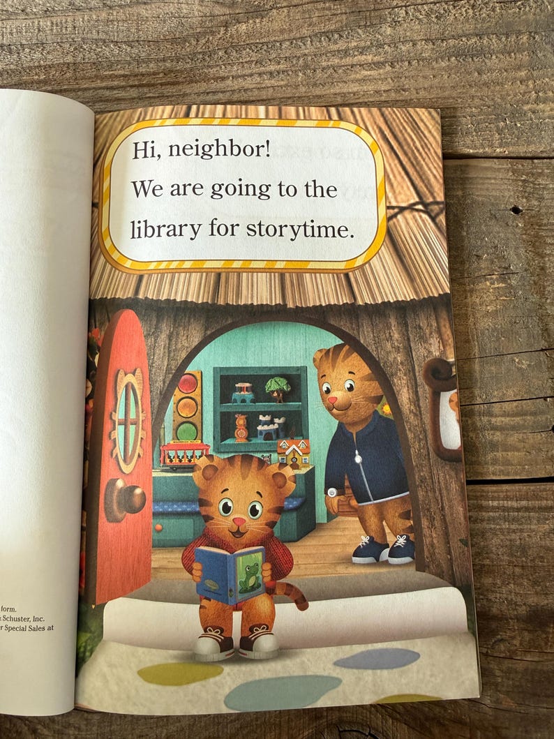 May include: A children's book page with the text "Hi, neighbor! We are going to the library for storytime." The illustration features a tiger character holding a book and another tiger character standing in a doorway.
