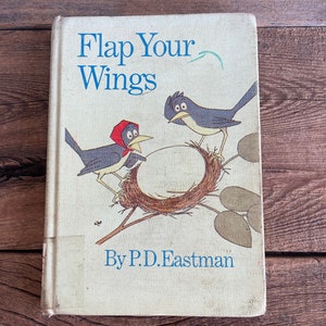 Könnte beinhalten: Ein altes Kinderbuch mit dem Titel "Flap Your Wings" von P.D. Eastman, mit einer Illustration von zwei Vögeln in einem Nest auf dem Cover.