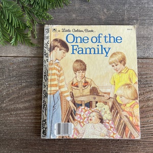 Pu&ograve; includere: Un libro vintage della serie Little Golden Book intitolato "One of the Family". La copertina presenta una famiglia di bambini, tra cui un bambino in una culla, con una combinazione di colori giallo, marrone e blu.