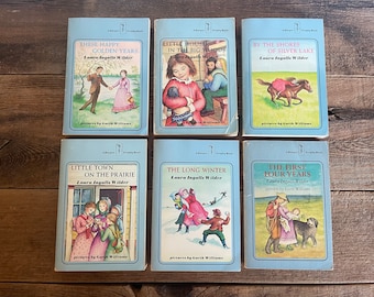 May include: A set of six blue paperback books with white text and illustrations. The books are part of the "Little House" series by Laura Ingalls Wilder. The titles are "These Happy Golden Years", "Little House in the Big Woods", "By the Shores of Silver Lake", "Little Town on the Prairie", "The Long Winter", and "The First Four Years".