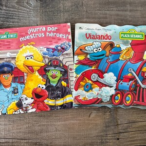 May include: Two Sesame Street children's books. One book cover features Big Bird, Elmo, a Dalmatian, and a police officer and firefighter. The other book cover shows a train with Cookie Monster and the text "Viajando".