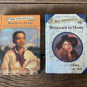 Puede incluir: Dos libros de bolsillo con cubiertas azules y naranjas. El libro de la izquierda se titula "My America: Message in the Sky, Corey's Underground Railroad Diary". El libro de la derecha se titula "My America: Westward to Home, Olivia's Diary, The Oregon Trail, 1848."