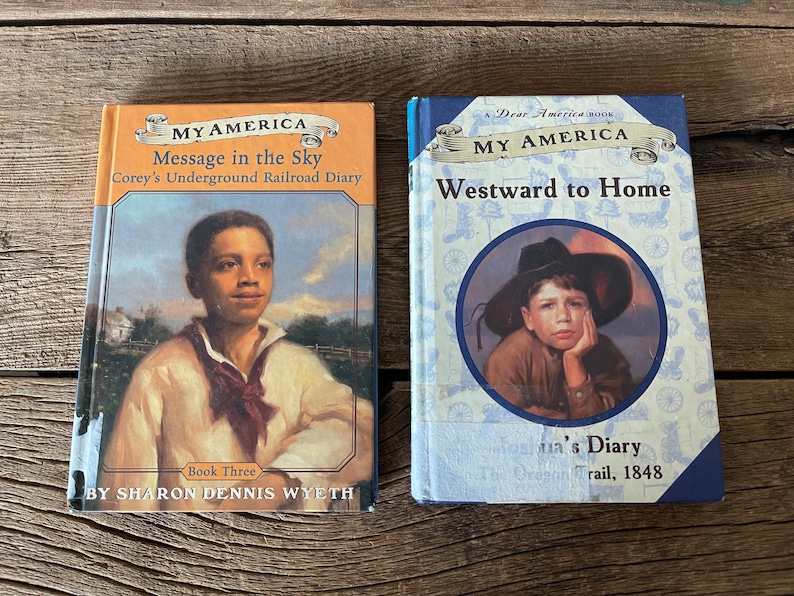 Puede incluir: Dos libros de bolsillo con cubiertas azules y naranjas. El libro de la izquierda se titula "My America: Message in the Sky, Corey's Underground Railroad Diary". El libro de la derecha se titula "My America: Westward to Home, Olivia's Diary, The Oregon Trail, 1848."