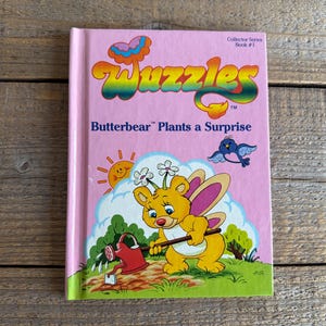 Könnte beinhalten: Ein Vintage-Kinderbuch mit dem Titel "Wuzzles Butterbear Plants a Surprise". Das Cover ist rosa mit farbenfrohen Illustrationen von Zeichentrickfiguren, darunter ein gelber Bär mit Schmetterlingsflügeln, eine Gießkanne und ein blauer Vogel. Das Buch gehört zur Collector-Serie.