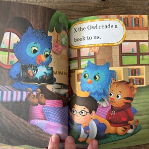 May include: An open book illustration featuring a blue owl reading to other cartoon characters. The text "X the Owl reads a book to us" is displayed. The scene includes a tree, pillows, and a bookshelf.