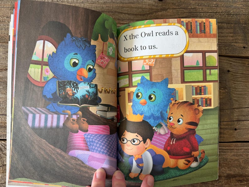 May include: An open book illustration featuring a blue owl reading to other cartoon characters. The text "X the Owl reads a book to us" is displayed. The scene includes a tree, pillows, and a bookshelf.