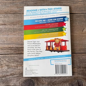 May include: Back cover of a children's book titled "Daniel Tiger and Prince Wednesday". The cover features a colorful design with text and an illustration of Daniel Tiger on a red trolley. The book is part of the "Reading with the Stars" series.