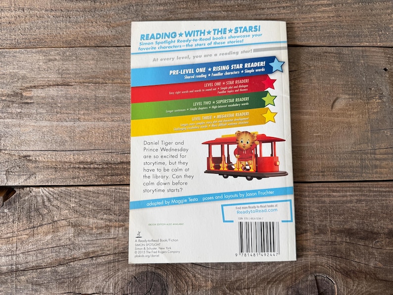 May include: Back cover of a children's book titled "Daniel Tiger and Prince Wednesday". The cover features a colorful design with text and an illustration of Daniel Tiger on a red trolley. The book is part of the "Reading with the Stars" series.