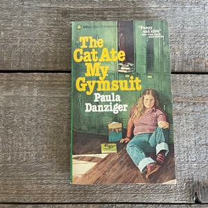 May include: A green paperback book with the title "The Cat Ate My Gymsuit" by Paula Danziger. The cover features a young girl sitting on the floor in front of a row of green lockers.