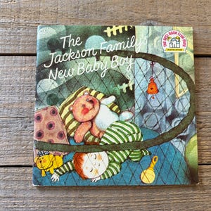 Pode incluir: Um livro infantil vintage intitulado "The Jackson Family's New Baby Boy". A capa apresenta uma ilustração de um bebê com um ursinho de pelúcia, um sino e um brinquedo amarelo. O livro é do The Best Book Club.