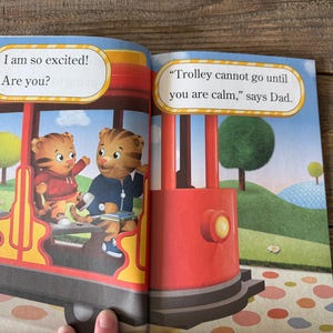 May include: A children's book spread featuring illustrations of Daniel Tiger and a friend on a red trolley. Text bubbles read "I am so excited! Are you?" and "Trolley cannot go until you are calm," says Dad."