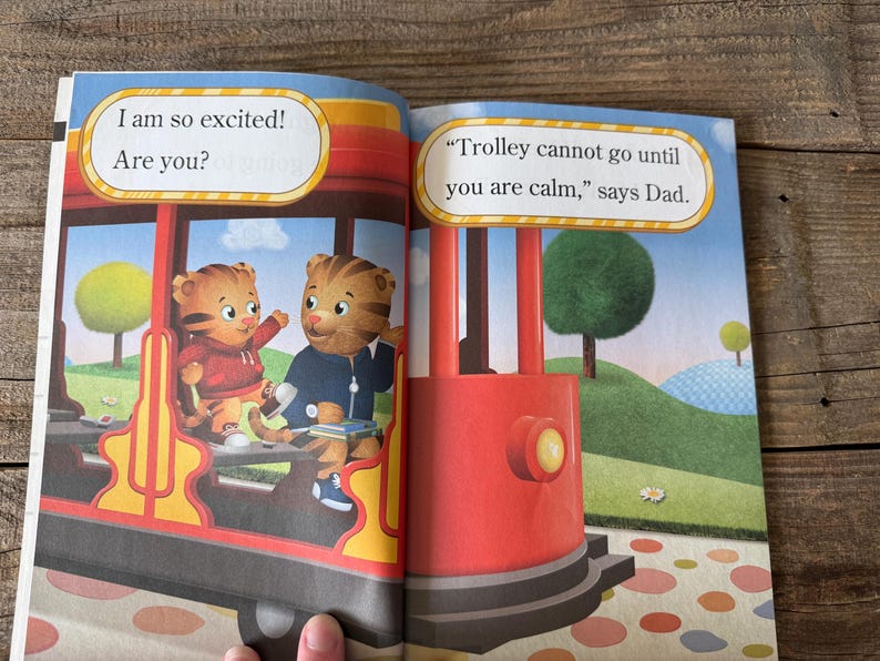 May include: A children's book spread featuring illustrations of Daniel Tiger and a friend on a red trolley. Text bubbles read "I am so excited! Are you?" and "Trolley cannot go until you are calm," says Dad."