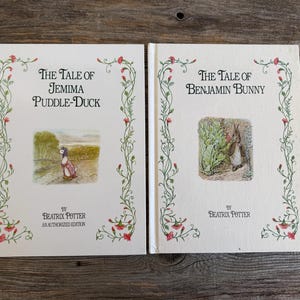 May include: Two vintage Beatrix Potter books, "The Tale of Jemima Puddle-Duck" and "The Tale of Benjamin Bunny," are displayed. The covers are cream-colored with floral and vine borders. The titles are in elegant black script, with illustrations of the characters.