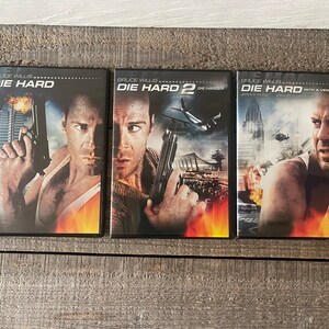 K&ouml;nnte beinhalten: Drei DVD-H&uuml;llen f&uuml;r die Die Hard-Filmreihe. Die H&uuml;llen sind schwarz mit rotem und orangefarbenem Hintergrund. Die erste H&uuml;lle zeigt Bruce Willis mit einer Waffe in der Hand und dem Text "Die Hard". Die zweite H&uuml;lle zeigt Bruce Willis mit einer Waffe in der Hand und dem Text "Die Hard 2 Die Harder". Die dritte H&uuml;lle zeigt Bruce Willis mit einer Waffe in der Hand und dem Text "Die Hard With a Vengeance".