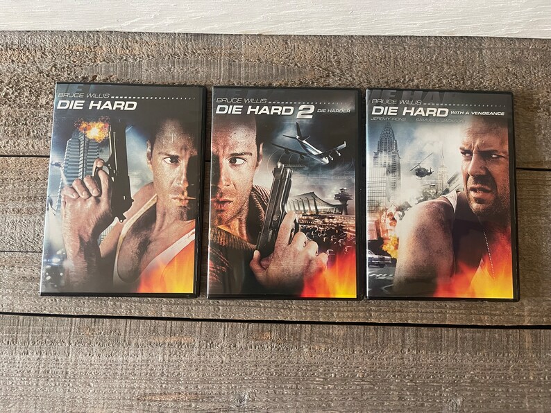 K&ouml;nnte beinhalten: Drei DVD-H&uuml;llen f&uuml;r die Die Hard-Filmreihe. Die H&uuml;llen sind schwarz mit rotem und orangefarbenem Hintergrund. Die erste H&uuml;lle zeigt Bruce Willis mit einer Waffe in der Hand und dem Text "Die Hard". Die zweite H&uuml;lle zeigt Bruce Willis mit einer Waffe in der Hand und dem Text "Die Hard 2 Die Harder". Die dritte H&uuml;lle zeigt Bruce Willis mit einer Waffe in der Hand und dem Text "Die Hard With a Vengeance".