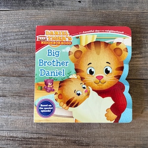 Puede incluir: Un libro infantil titulado "Big Brother Daniel" de Daniel Tiger's Neighborhood. La portada presenta una ilustraci&oacute;n de Daniel Tiger sosteniendo a otro cachorro de tigre. El libro tiene un borde rojo y azul y est&aacute; sobre un fondo de madera.