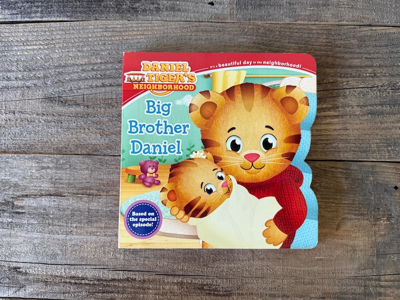 Puede incluir: Un libro infantil titulado "Big Brother Daniel" de Daniel Tiger's Neighborhood. La portada presenta una ilustraci&oacute;n de Daniel Tiger sosteniendo a otro cachorro de tigre. El libro tiene un borde rojo y azul y est&aacute; sobre un fondo de madera.