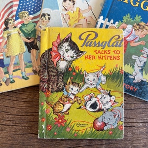 May include: A yellow children's book titled "Pussy Cat Talks to Her Kittens" featuring illustrations of a cat and kittens playing with string. The book is surrounded by other children's books with illustrations of children and animals.