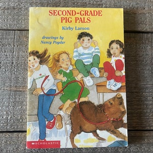 Op de afbeelding: Een kinderboek getiteld "Second-Grade Pig Pals" van Kirby Larson met illustraties van Nancy Poydar. De omslag toont vier kinderen en een varken in een klaslokaal.