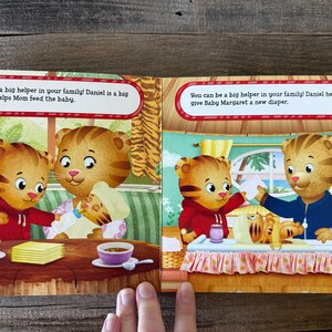 Puede incluir: Un libro infantil abierto en dos p&aacute;ginas con ilustraciones de Daniel el Tigre. El texto dice: "&iexcl;Puedes ser una gran ayuda en tu familia! Daniel es una gran ayuda. &Eacute;l ayuda a mam&aacute; a alimentar al beb&eacute;." y "&iexcl;Puedes ser una gran ayuda en tu familia! Daniel ayuda a pap&aacute; a darle a Baby Margaret un pa&ntilde;al nuevo."