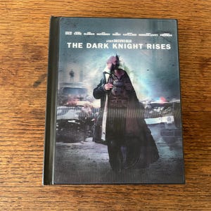 Puede incluir: Caja de DVD de la película "The Dark Knight Rises". La portada presenta un personaje con abrigo oscuro y máscara frente a una escena de la ciudad, con el título de la película en la parte superior.