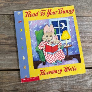 Puede incluir: Libro infantil titulado "Read To Your Bunny" de Rosemary Wells. La portada presenta un conejo leyendo a otro conejo más pequeño en un sillón verde. El fondo muestra una ventana con una luna y una lámpara amarilla.