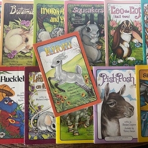 May include: A collection of 11 children's books with colorful covers featuring various animals, including a bear, a rabbit, a deer, a squirrel, a bunny, a fly, a bug, a mouse, a donkey, and a horse. The book titles include "Jingle Bear", "Buttermilk", "Morgan and You", "Squeakers", "Leo the Lop (tail two)", "Flutterby Fly", "Nikini", "Huckleberry", "Little Mouse on the Hill", "KiYomi", "Pish Posh", and "Shimmering".