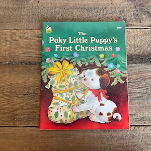 May include: A Golden Book titled "The Poky Little Puppy's First Christmas" featuring a puppy with brown spots and a red bow, holding a green and yellow wrapped gift under a Christmas tree with red and green ornaments.