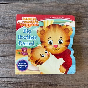 Puede incluir: Un libro infantil titulado "Big Brother Daniel" de Daniel Tiger's Neighborhood. La portada presenta una ilustraci&oacute;n de Daniel Tiger sosteniendo un beb&eacute; tigre. El libro tiene un borde rojo y azul y el texto "Based on the special episode!"